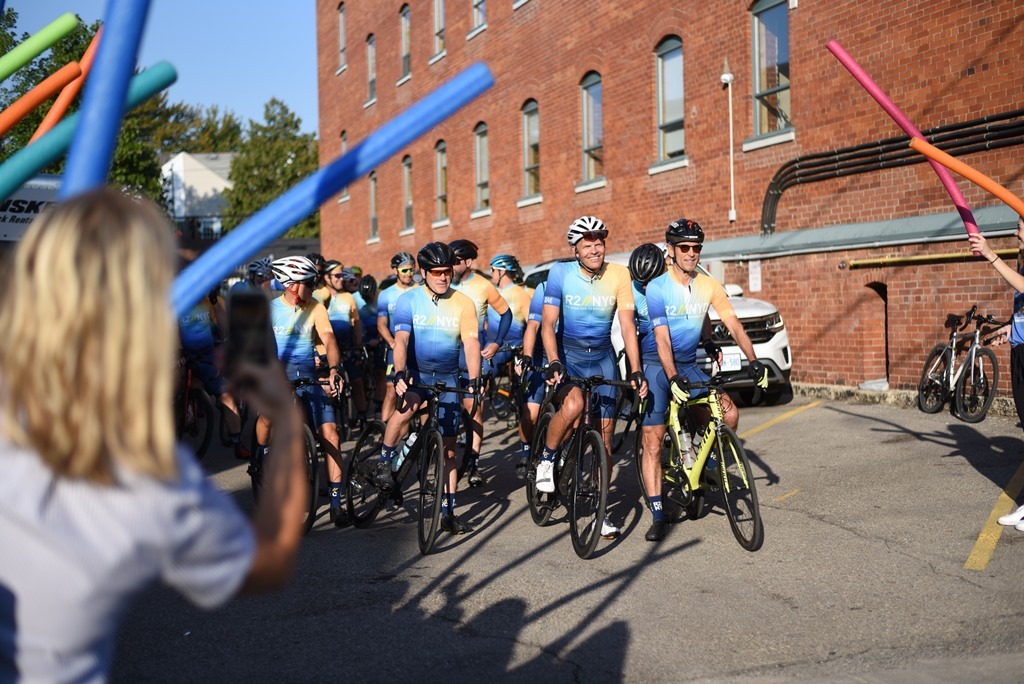 cyclists preparing to leave from parking lot with supporters cheering and holding pool noodles