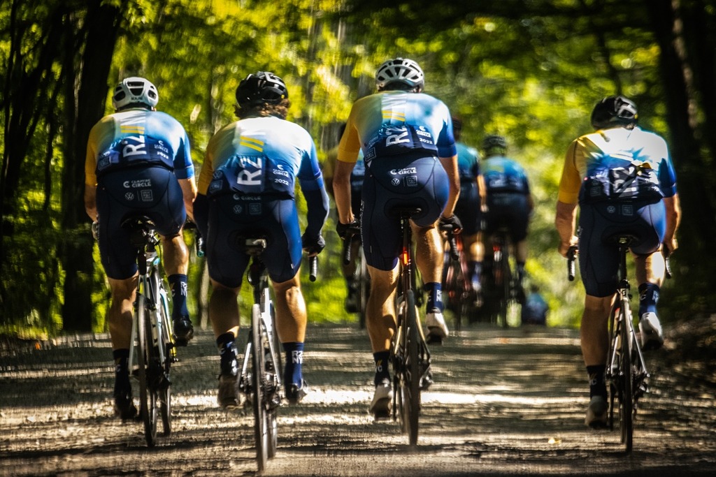 group of cyclists pictured riding down a scenic road surrounded by trees from behind