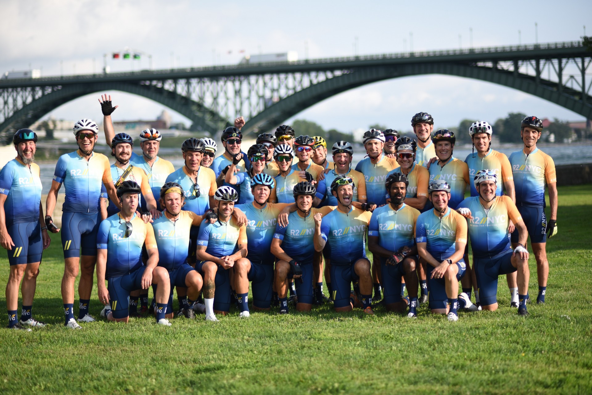 group picture of R2//NYC riders on a field with a bridge behind them